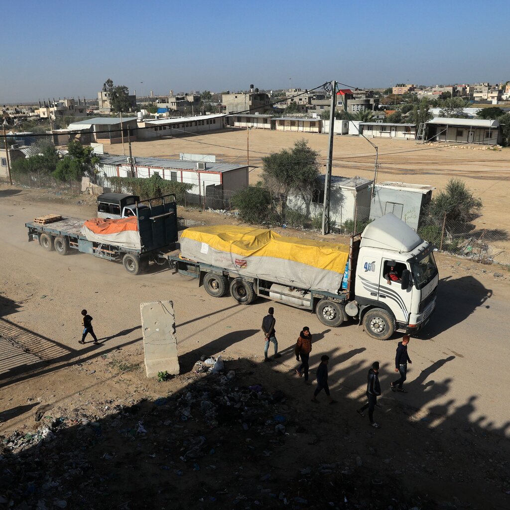 A truck with two beds, one covered in a yellow-and-gray covering and another with an orange-and-gray covering drives along a dusty road near a town. 