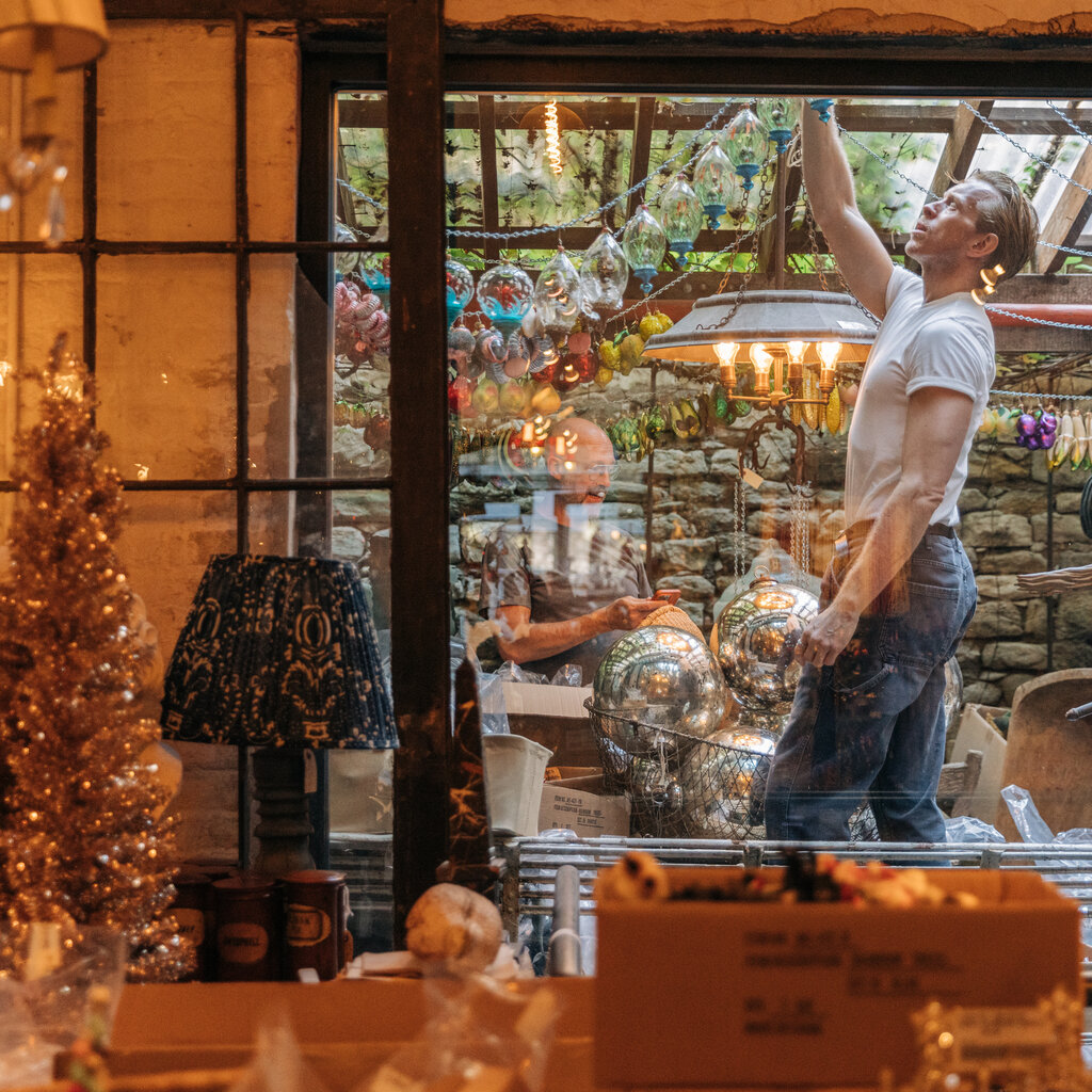 A man, seen through the window of a decorated store, arranging a string of ornaments. He is wearing a white T-shirt and jeans.