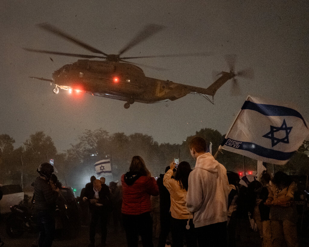 A military helicopter lands at night, with a crowd of people waiting below. 