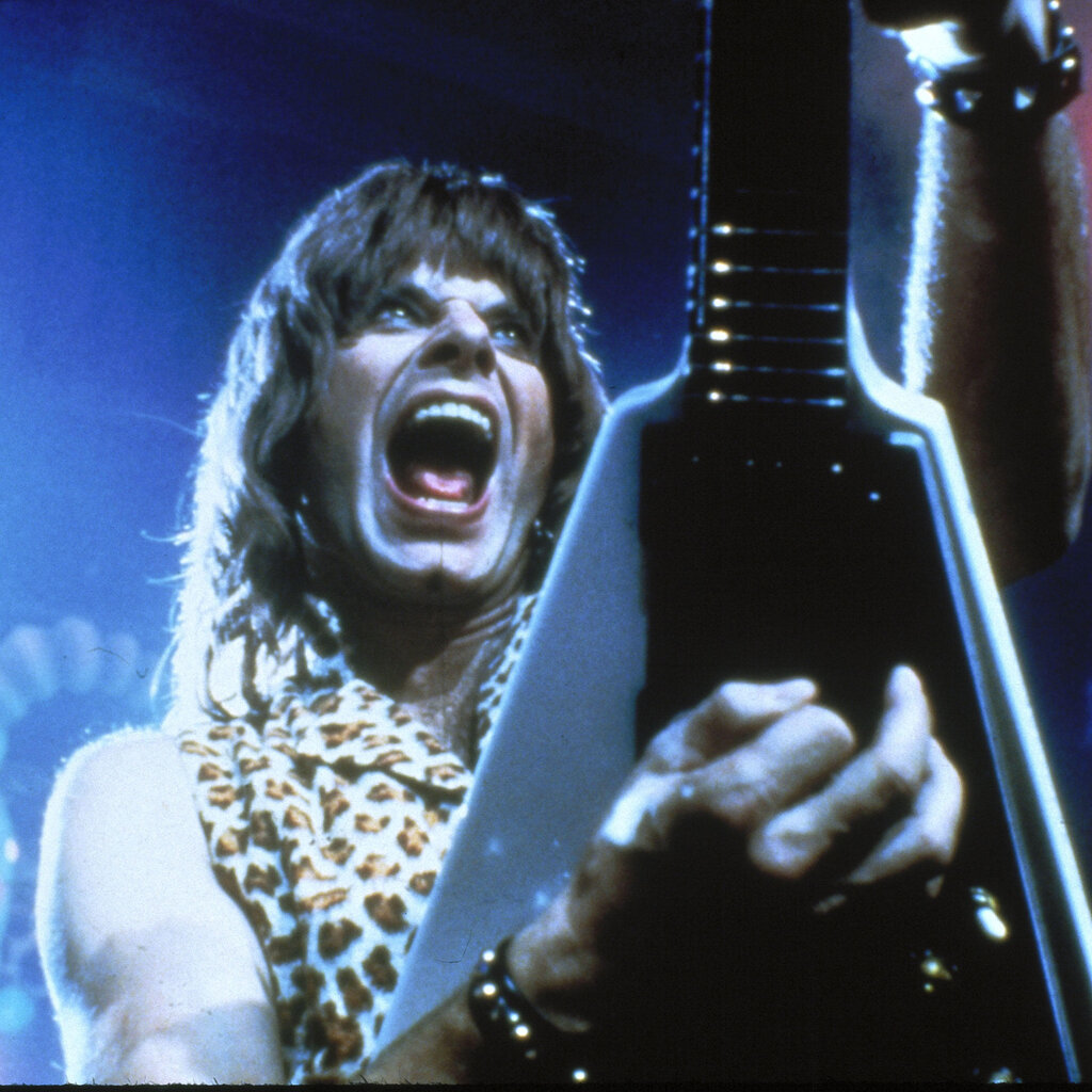 A man with long hair and bangs wears a leopard-print sleeveless top. He’s holding a triangle-shaped guitar upright.
