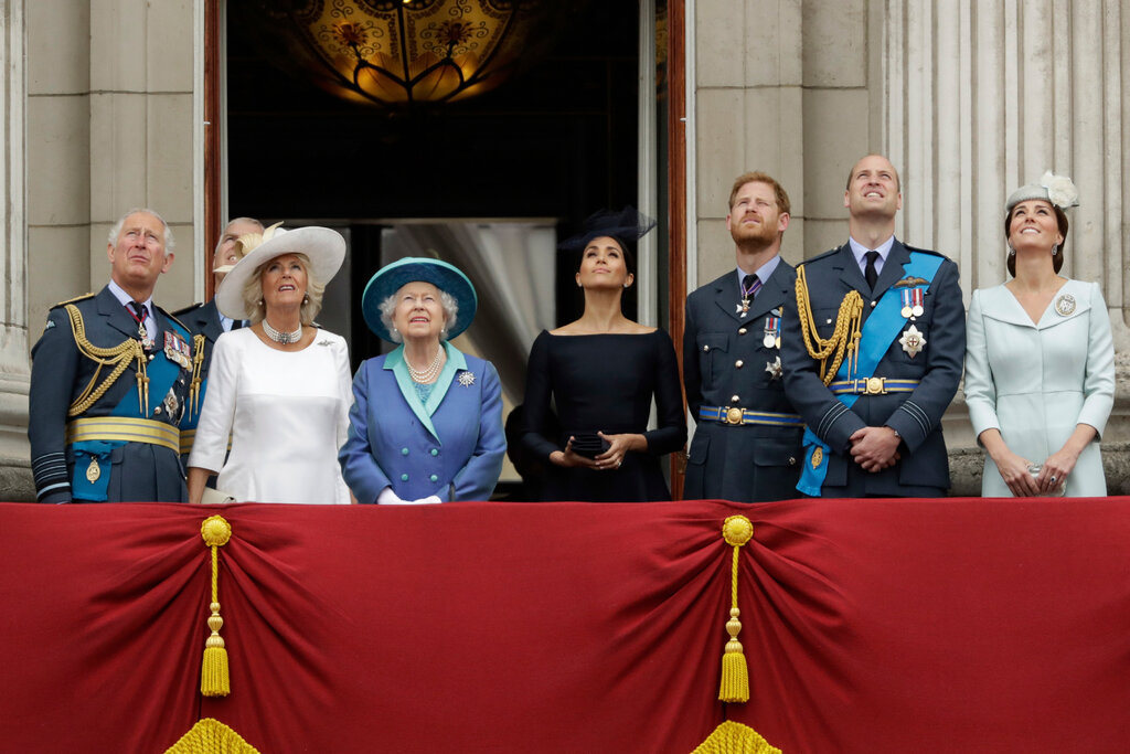 The Windsors gather on a red, draped balcony, looking upward. From left: Prince Charles in military regalia, Prince Andrew, Camilla the Duchess of Cornwall in a white dress and hat, Queen Elizabeth II in a blue dress and hat, Meghan the Duchess of Sussex in a black dress and fascinator, Prince Harry in military uniform, Prince William in military uniform and Catherine, the Duchess of Cambridge, in a pale-blue dress and pillbox hat. 