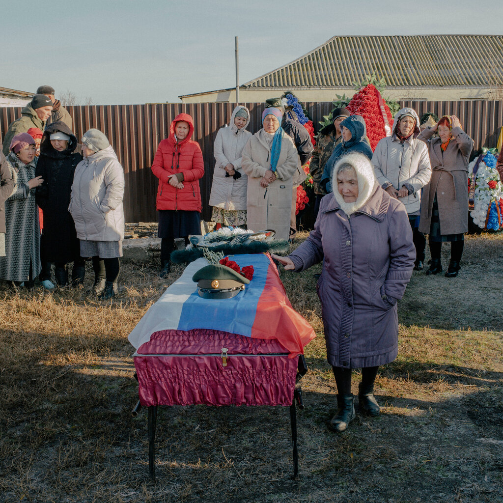 People wearing coats and hats surround a crimson coffin draped in a Russian flag. 