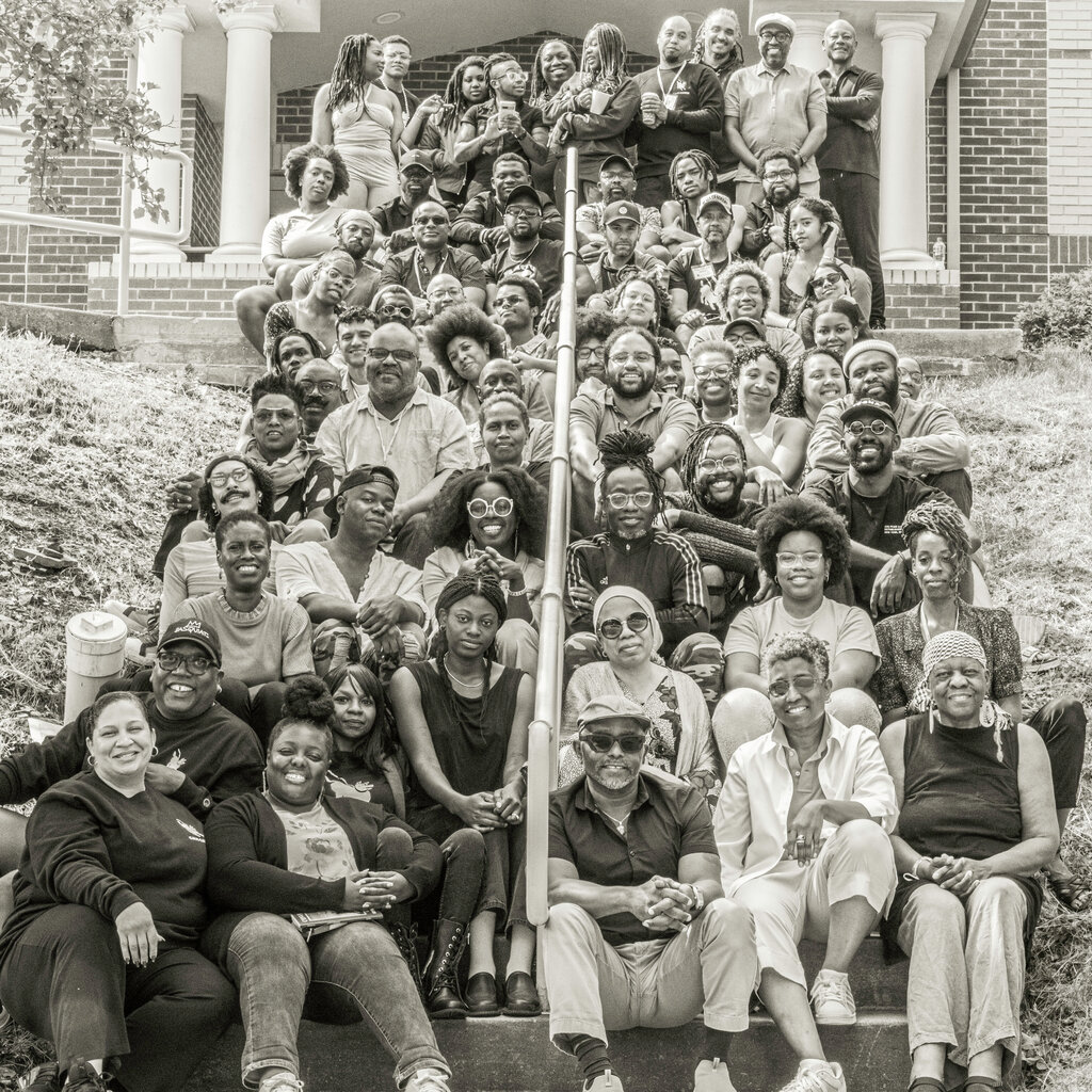 A black and white photograph of several dozen people sitting on the steps outside a brick building whose entrance is flanked by columns.