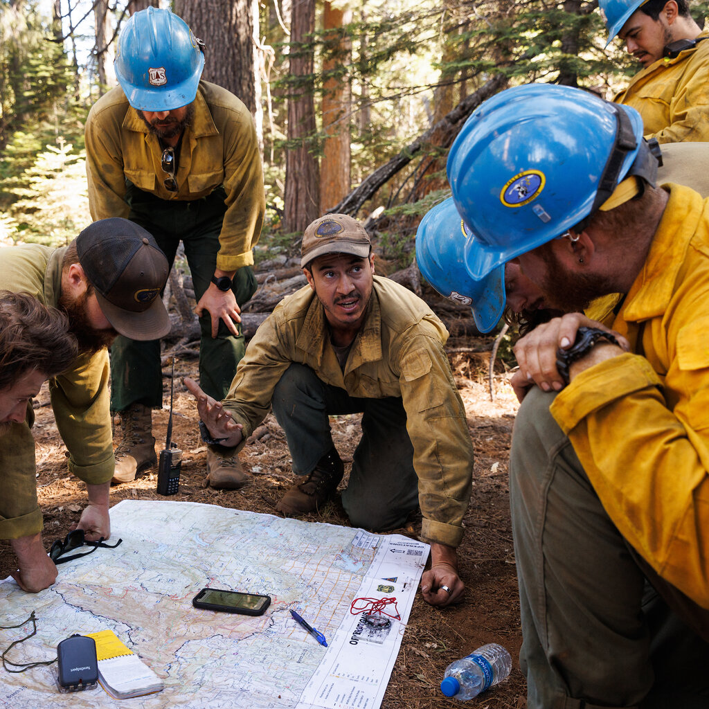 A group of firefighters kneeling over a map and one person speaking to the group.