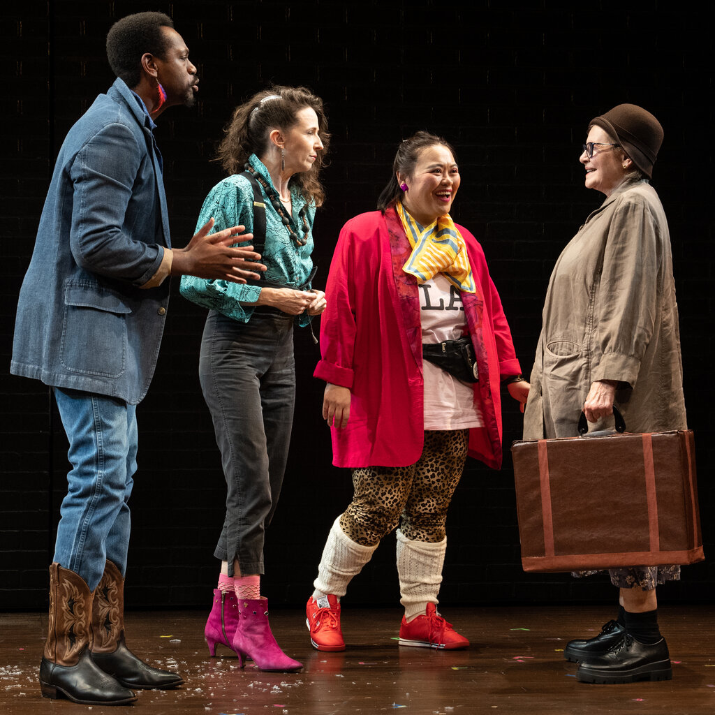 A man and two women look at a third woman in a raincoat who is holding a suitcase onstage,