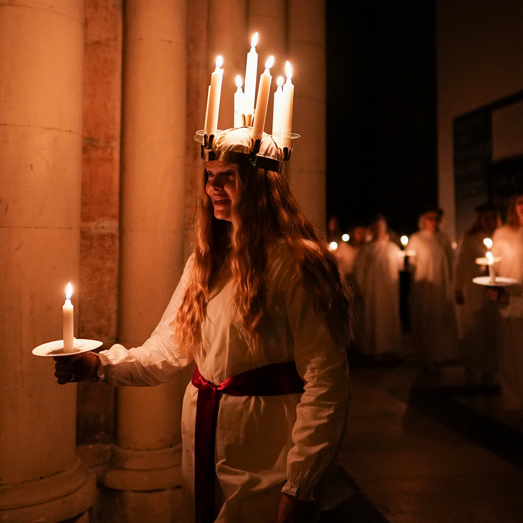 In a darkened room, which has large columns on one side, a procession of young women hold candles and wear white dresses with red sashes. The young woman in front has a crown-like adornment on her head, bedecked with candles.