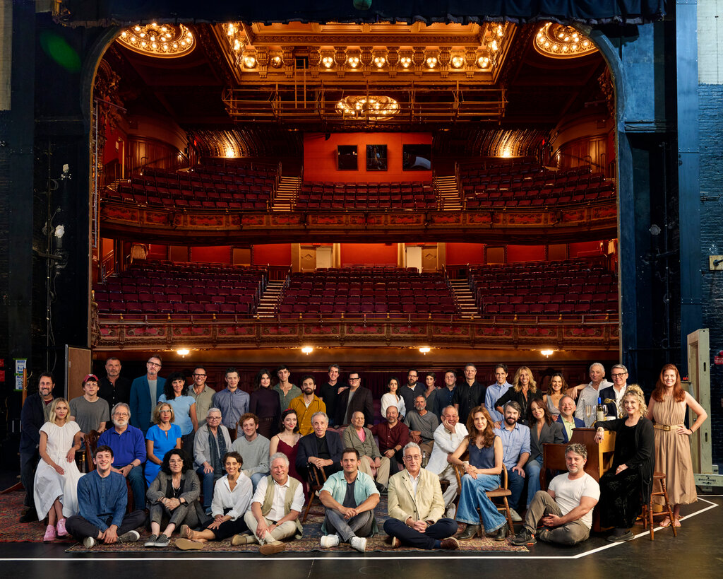 A group of about 50 people pose for a portrait on a Broadway stage, the camera looking out from the stage onto an empty audience of red velvet seats.