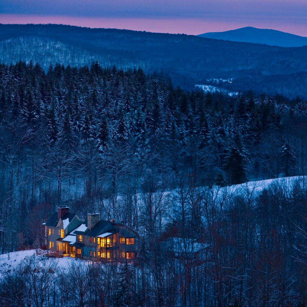 On a snowy hilltop at sunset, against a mountainous backdrop, a traditional house-like hotel with two chimneys is lit up invitingly.
