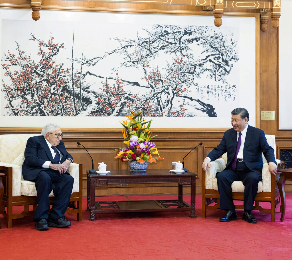Henry Kissinger and President Xi Jinping sitting in armchairs that flank a coffee table with a large bouquet in the middle. A brush painting hangs behind them. 