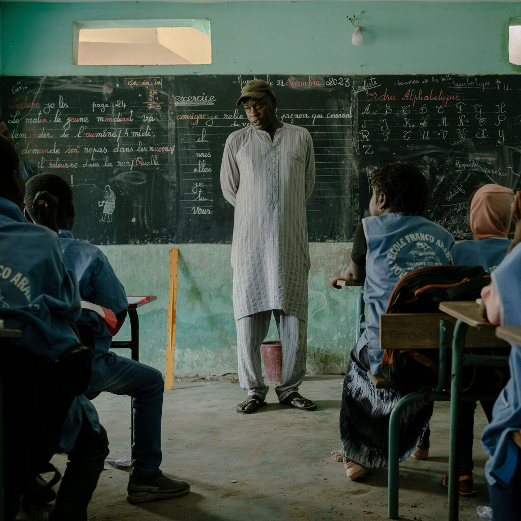 A man wearing a long shirt stands in front of a class of children, with a chalkboard behind him.