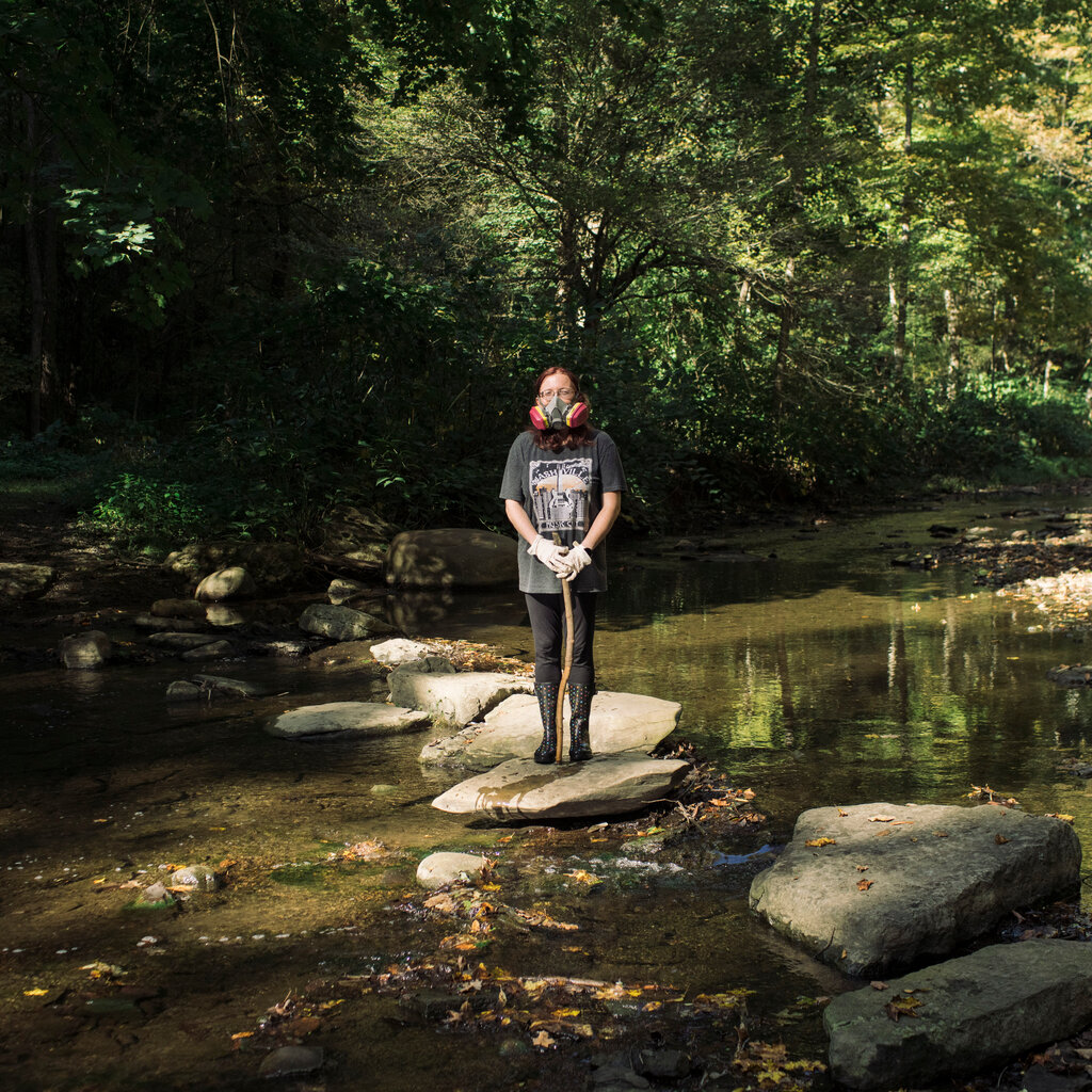A woman standing on a large rock in the middle of a creek surrounded by trees while wearing a respirator mask and gloves.