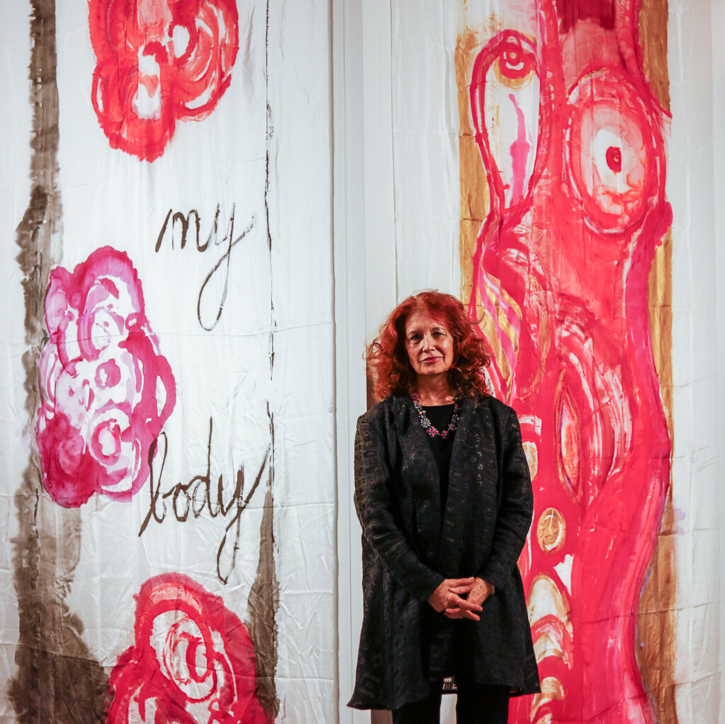 A woman stands against a backdrop of tall white walls with reddish art on them with the words “my body.”