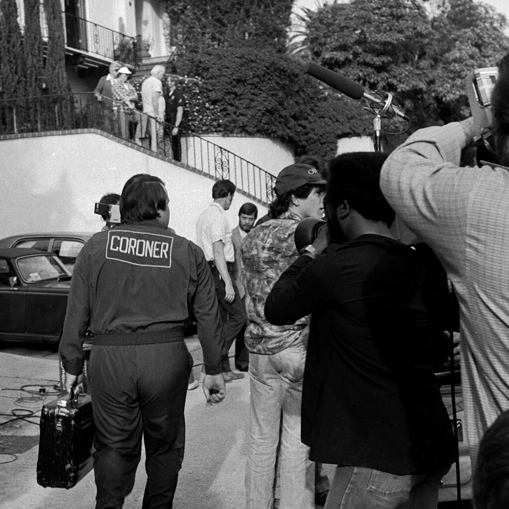 A black-and-white image of a man, carrying a case and wearing clothing that said “coroner” on the back, walking toward a staircase amid several members of the media. 