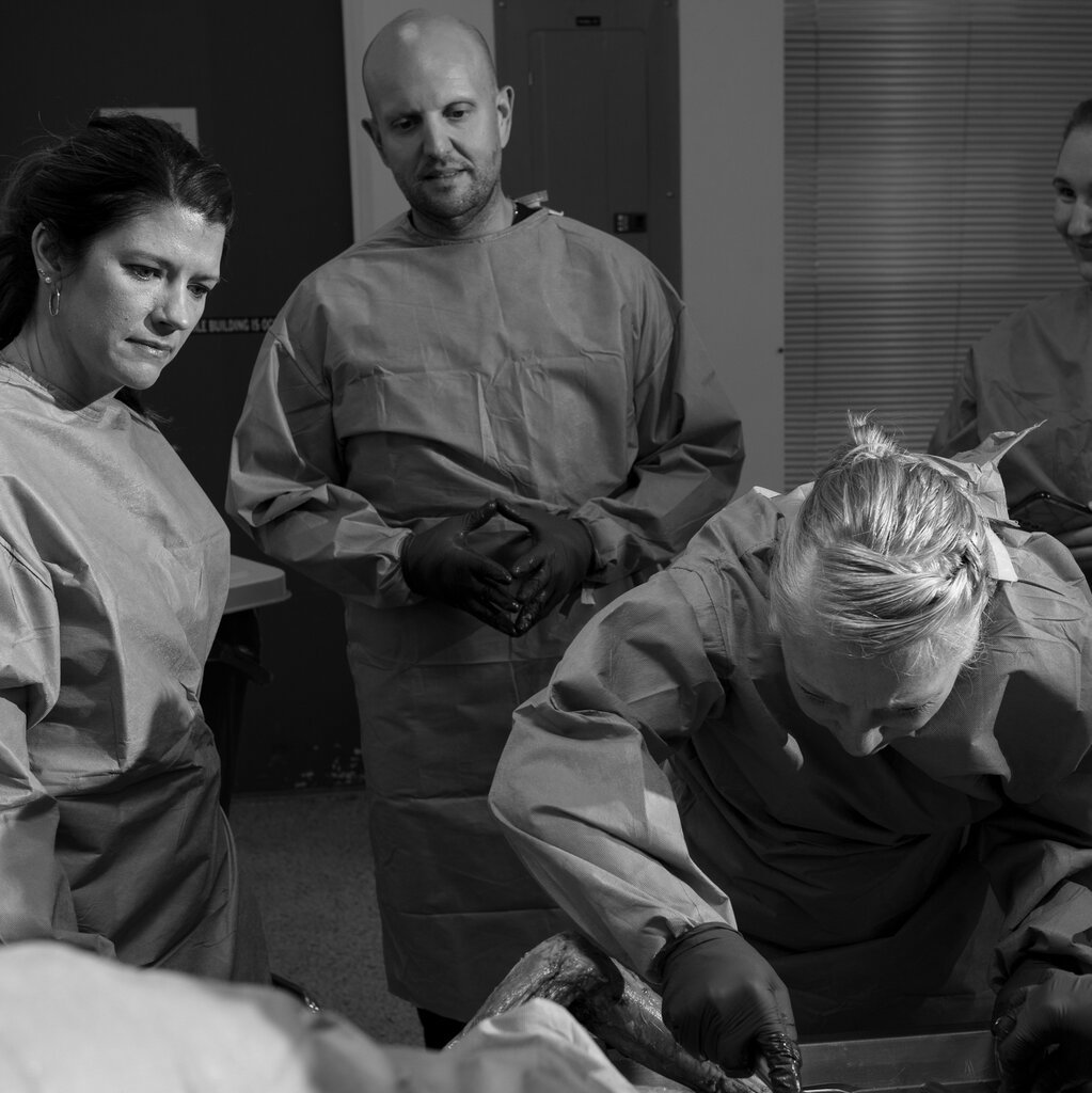 A black and white photo of a group of students wearing medical scrubs observing as one of them leans over to cut into a cadaver.