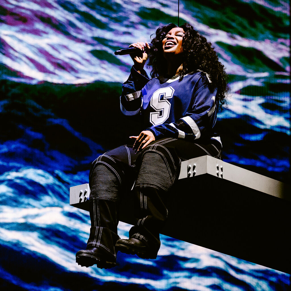SZA sings while sitting at the edge of a diving board with a screen showing the ocean behind her.