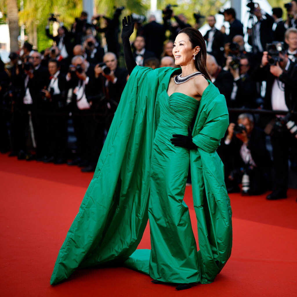 Michelle Yeoh waving on a red carpet. She is wearing a green gown with a matching stole and black gloves. 