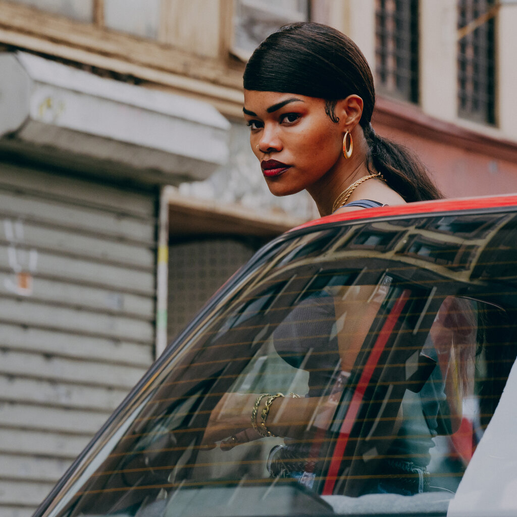 Teyana Taylor, black hair brushed low on her forehead and looking stern, is out of a car on a street with shuttered storefronts. 
