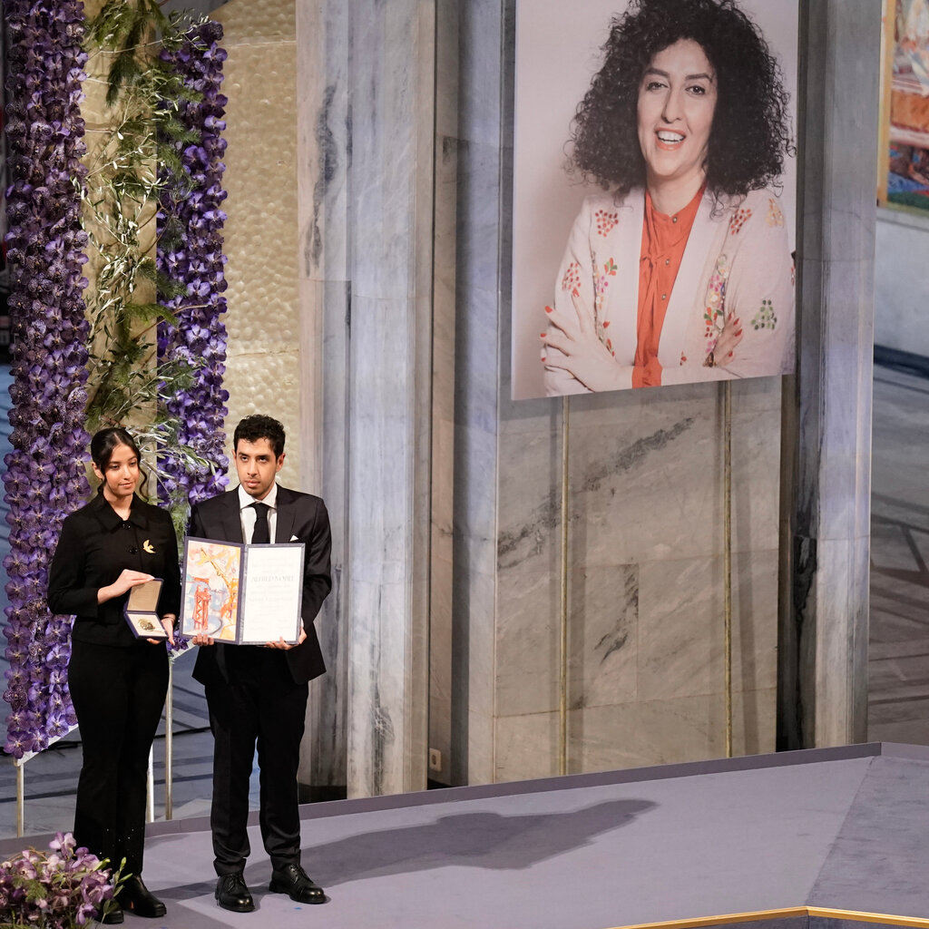 A young woman hold an open medal case, beside her a young man holds an open folder. They are on a stage. On the wall next to them is a photo of a woman with her arms folded, smiling. 