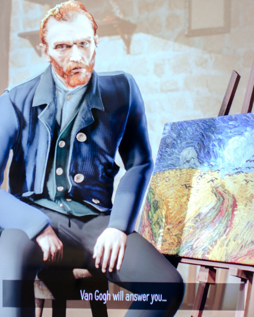 A replica of Vincent Van Gogh, through A.I., sits in a blue shirt, blue waistcoat and black pants, with a painting on the easel behind him.