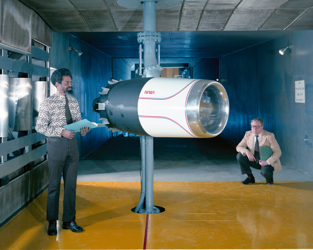 Two men in dress from the 1970s pose on either side of a jet engine that has the NASA worm logo on its side.