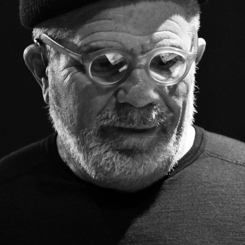 A black-and-white photograph of the playwright David Mamet, who is wearing thick-framed glasses and a dark T-shirt.