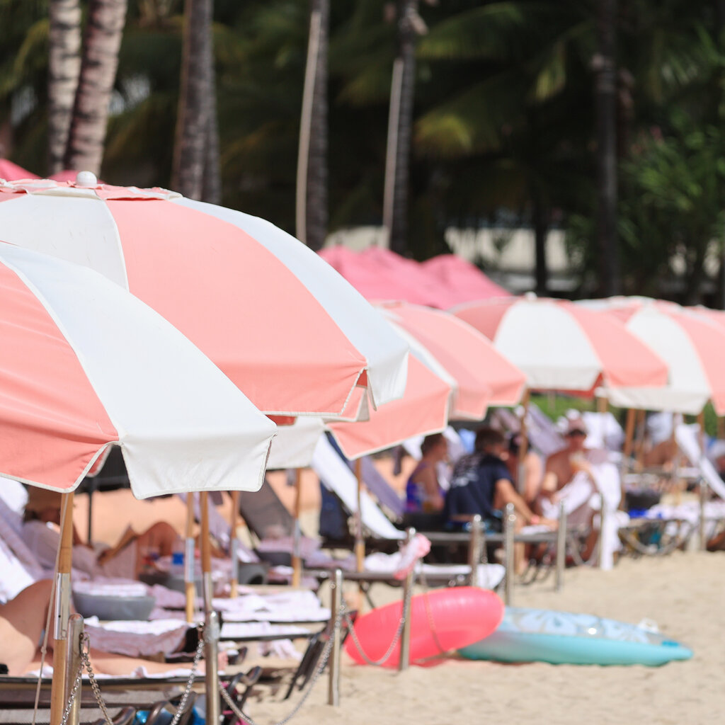 A line of pink-and-white beach umbrellas extends into the distance along a strip of sand. A few beach toys lie in front of them and people can be seen on chaise longues in the shade. 