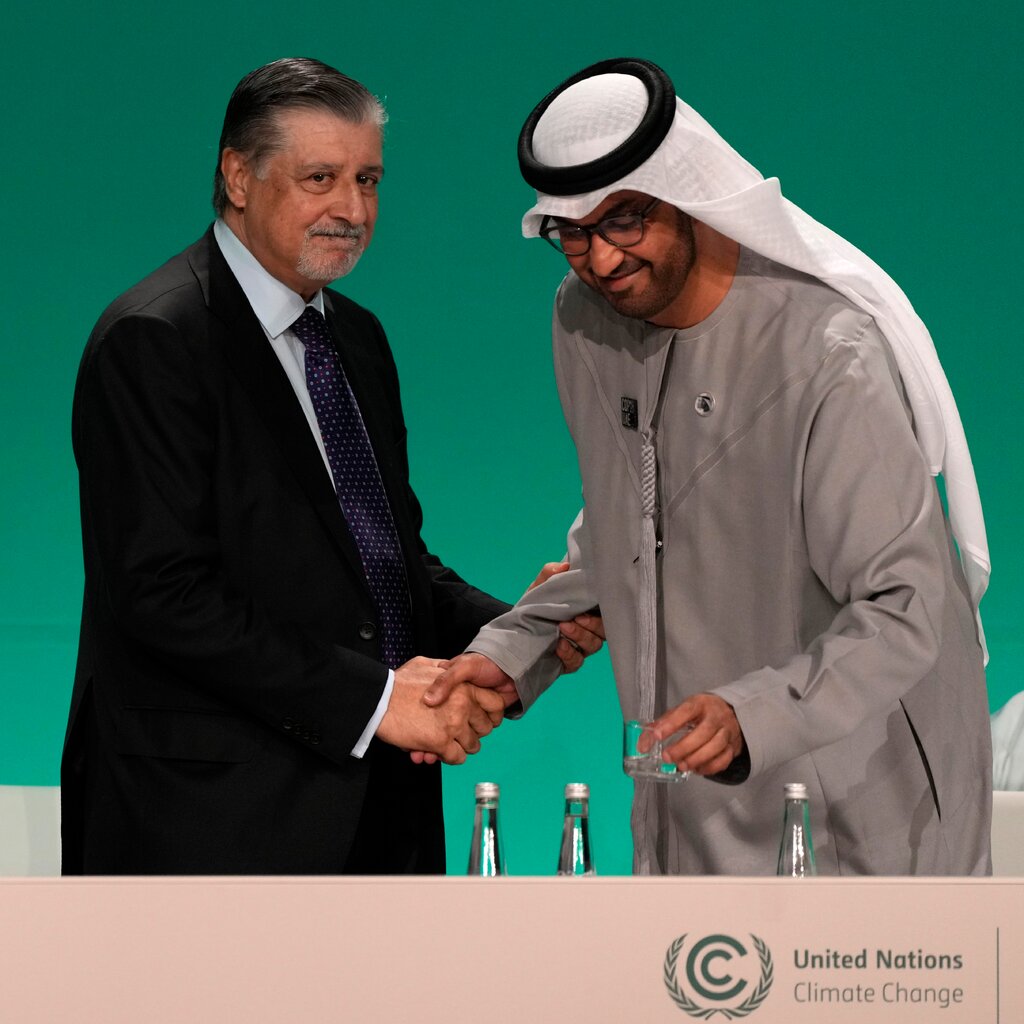 Three men on a stage with a green background. The two men on the right are shaking hands. 