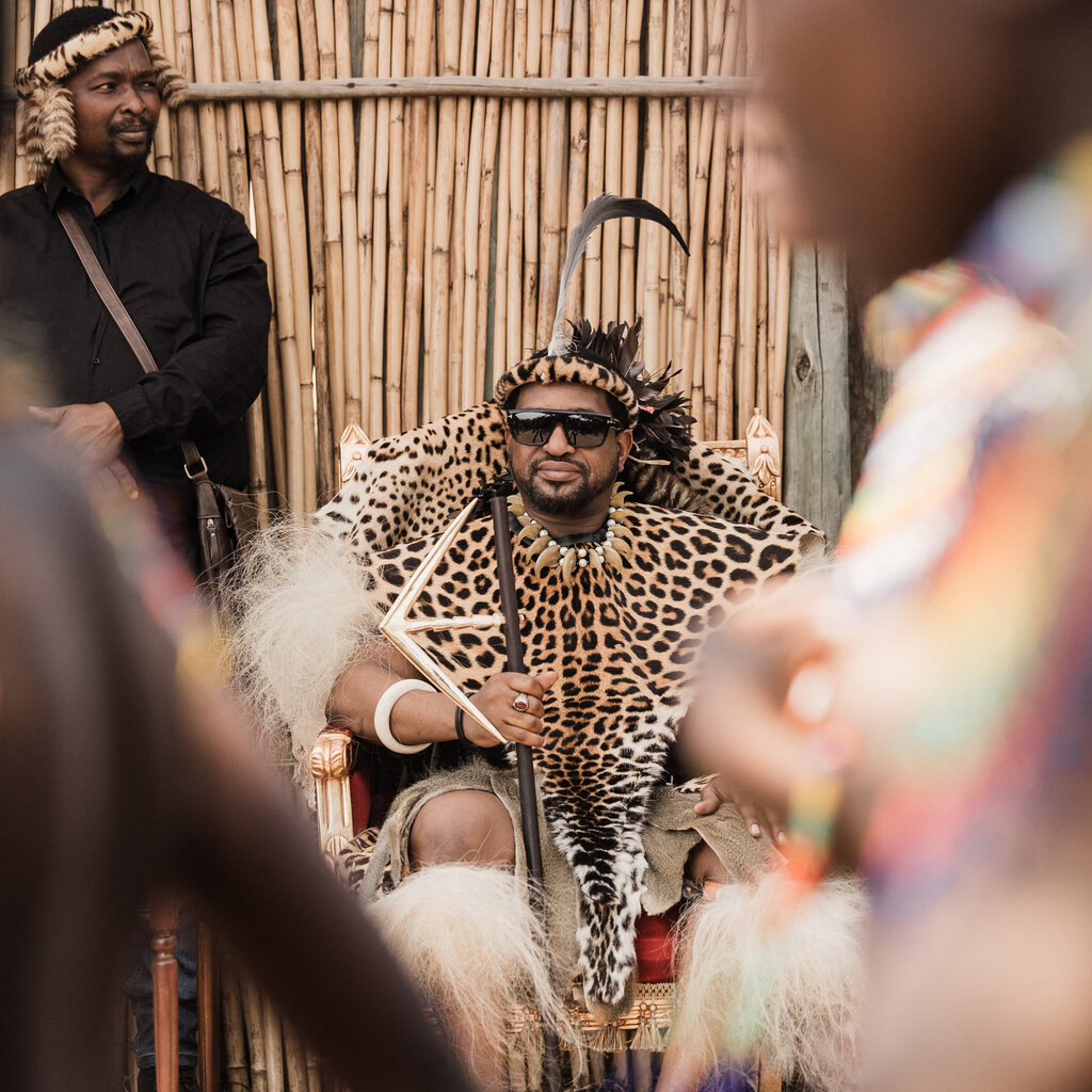 A Zulu king, wearing leopard print robes, sits on a throne. 