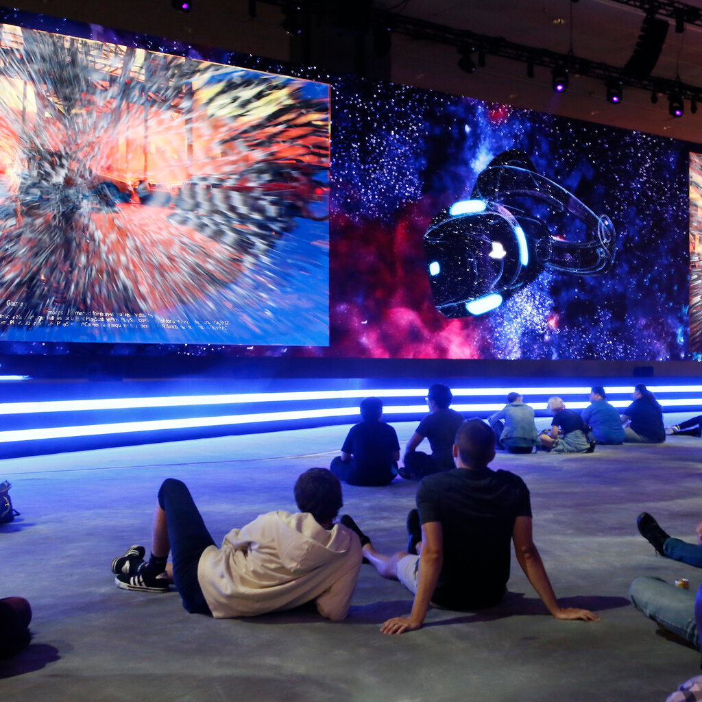 People sit on the ground in front of large screens. 