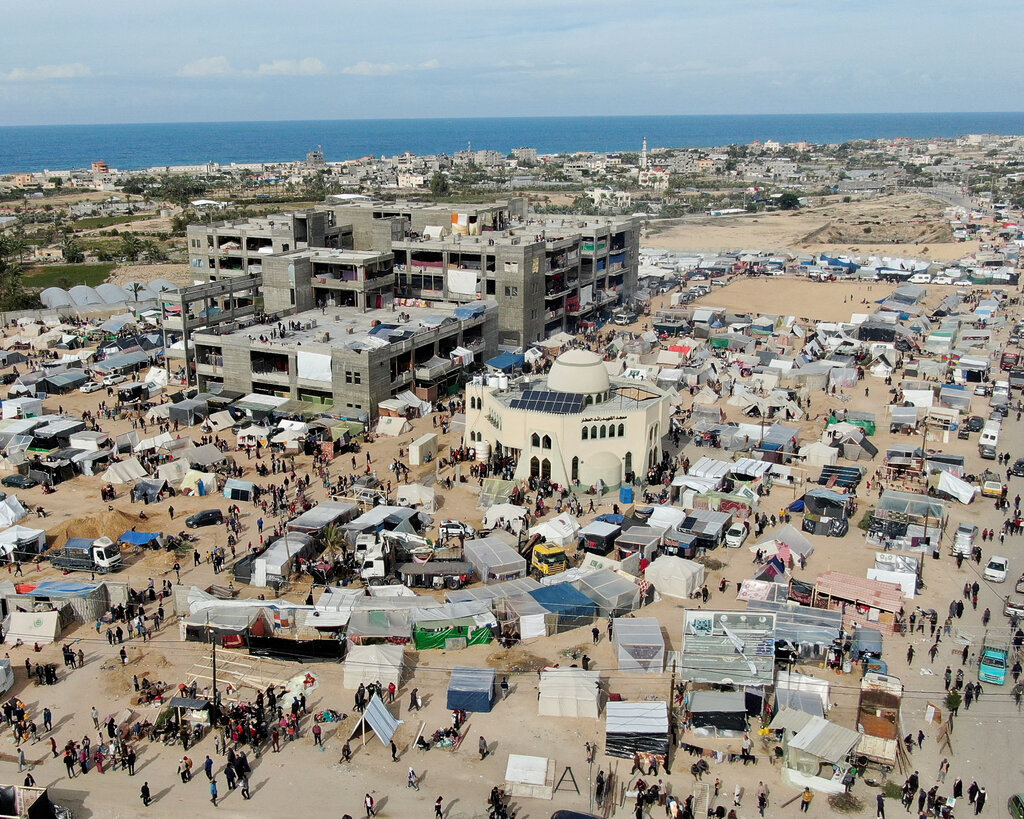 An aerial view showing a crowded complex of tents surrounding a mosque and a concrete building with the coastline and horizon in the background. 