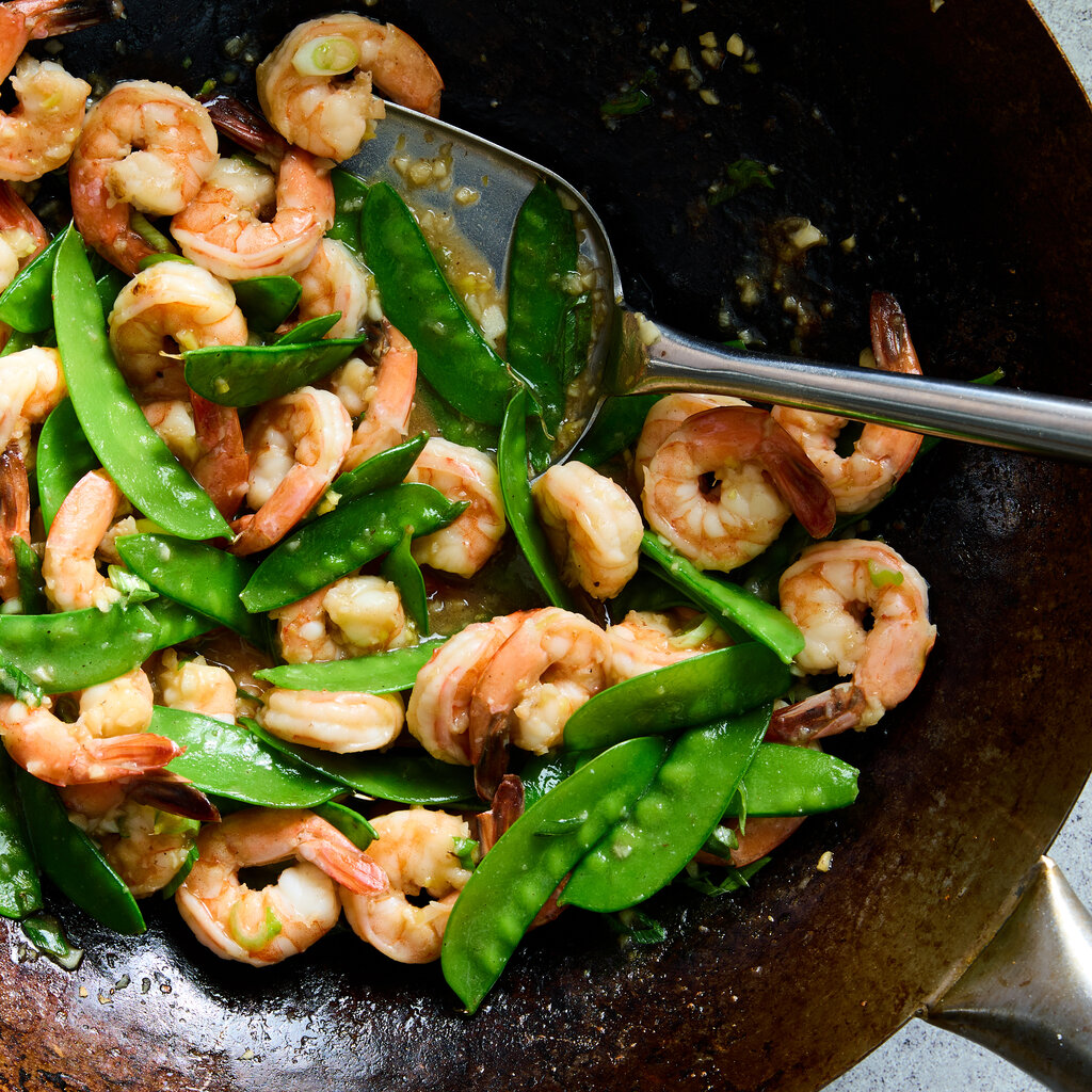 A wok holds stir-fried shrimp with snow peas and ginger with a wok spatula.