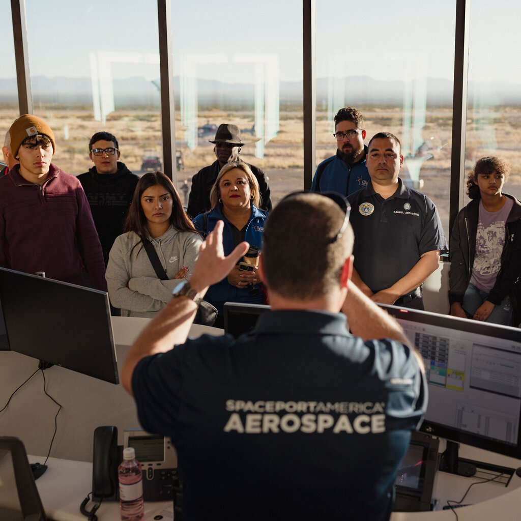 Allan Turk, seen from behind wearing a shirt with “Spaceport America Aerospace” written on the back, speaks across a table lined with computer monitors to a dozen people. They are standing in front of windows looking out on a desert.