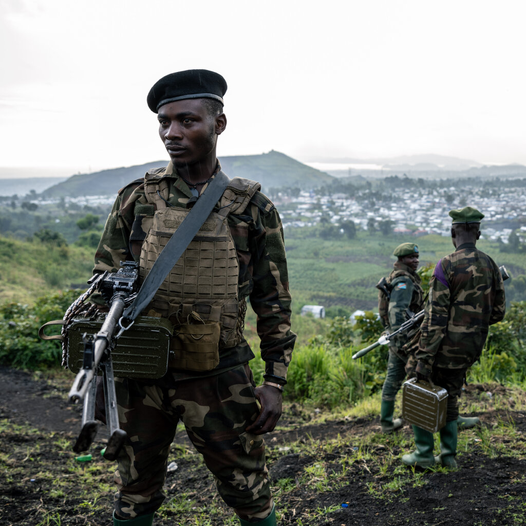 A soldier holding weaponry in the hills in the Democratic Republic of Congo. Other soldiers stand in the background. 