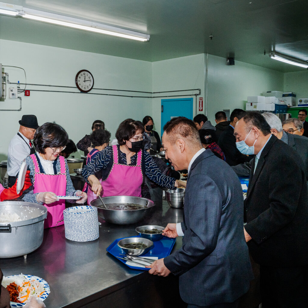 Women wearing bright pink aprons serve food from bowls. On the other side of a long table, men wearing suits hold trays. 