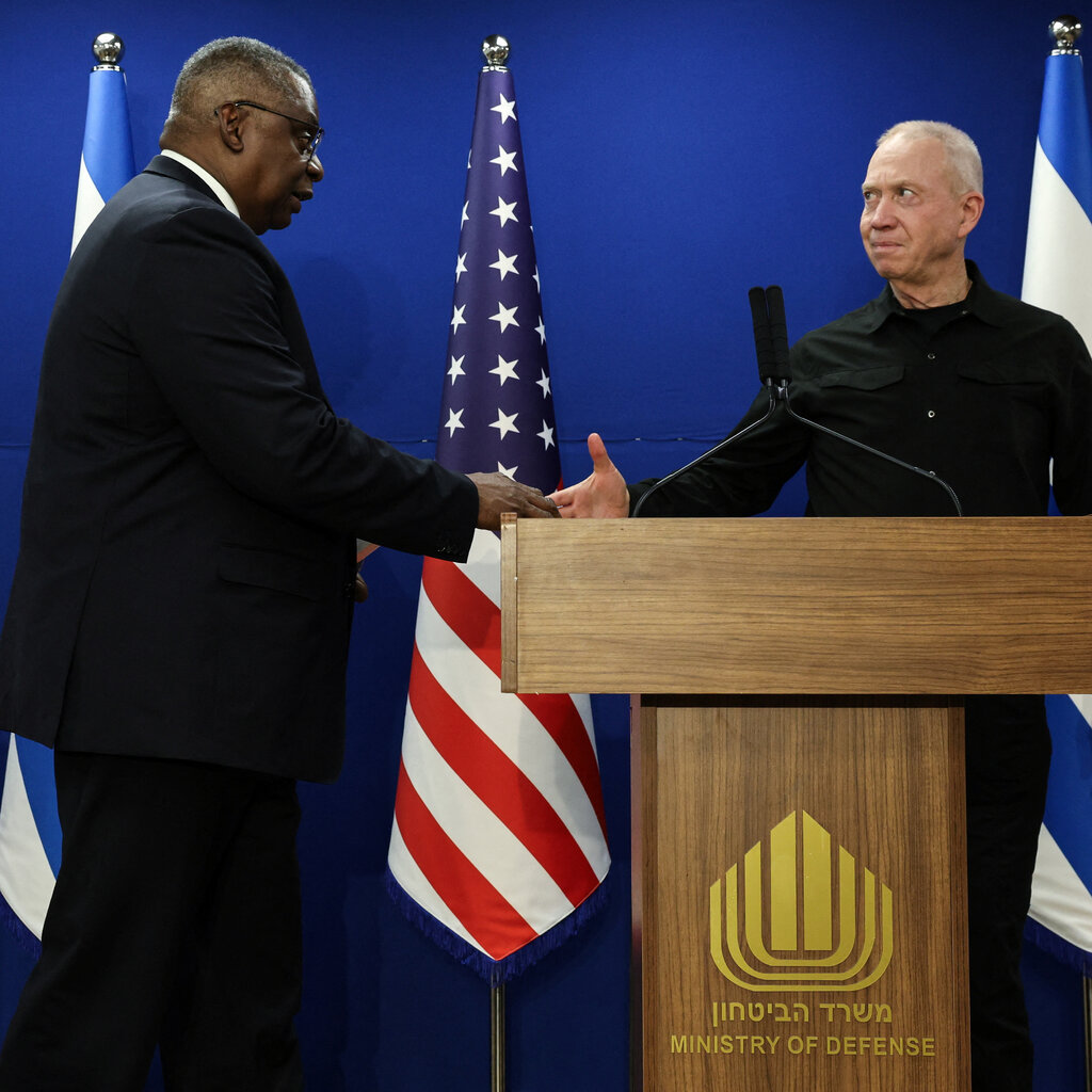 A man standing at a lectern with the words, "Ministry of Defense," wearing a black button-up shirt, looks to his right and shakes the hand of a man wearing a black suit. 
