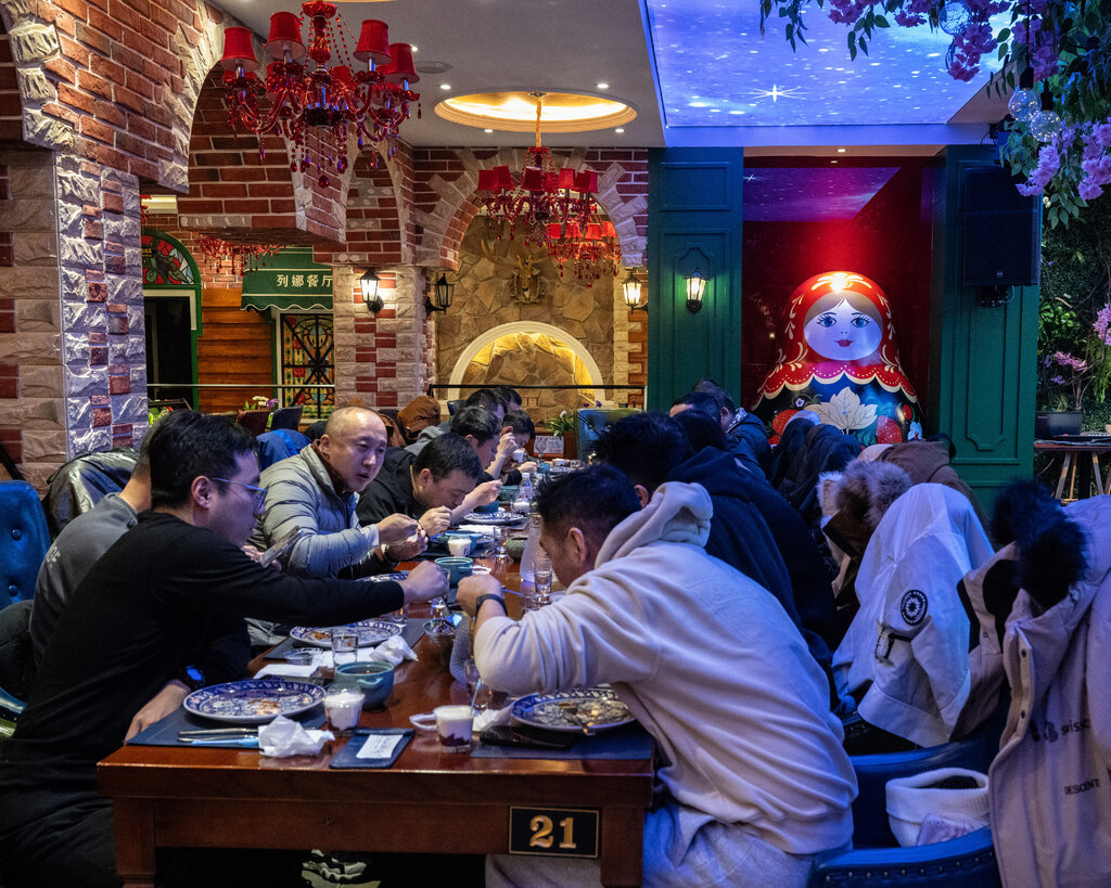 A group fills a long table at a restaurant. A large Russian nesting doll occupies a nook in the back.