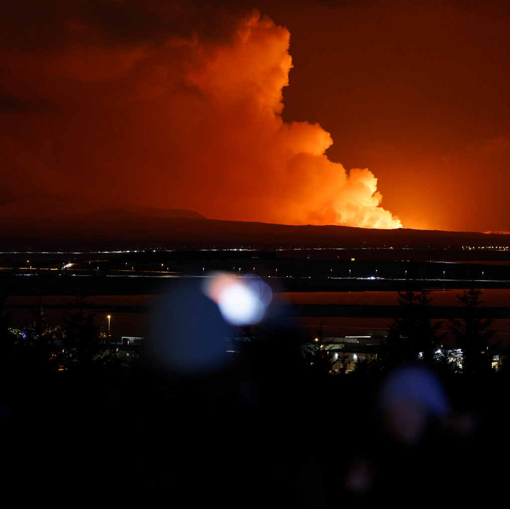 A view of smoke on the horizon, beyond city lights. The smoke is lit with a reddish-orange glow. 