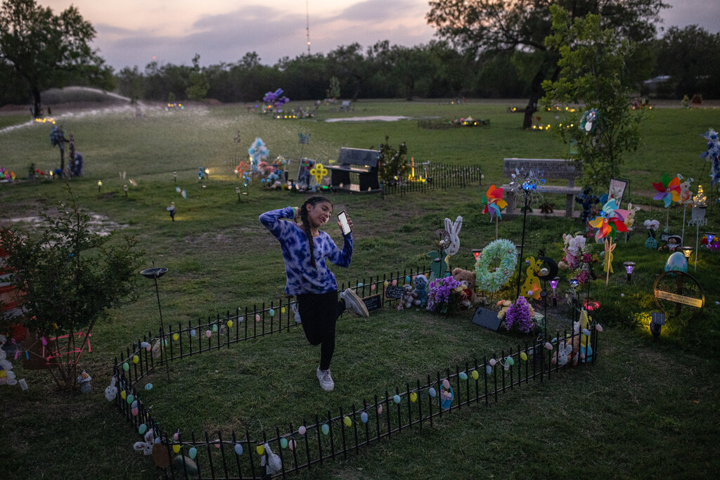 A girl in a purple sweater dances in a small, fenced off grave area, holding her phone near her ear.