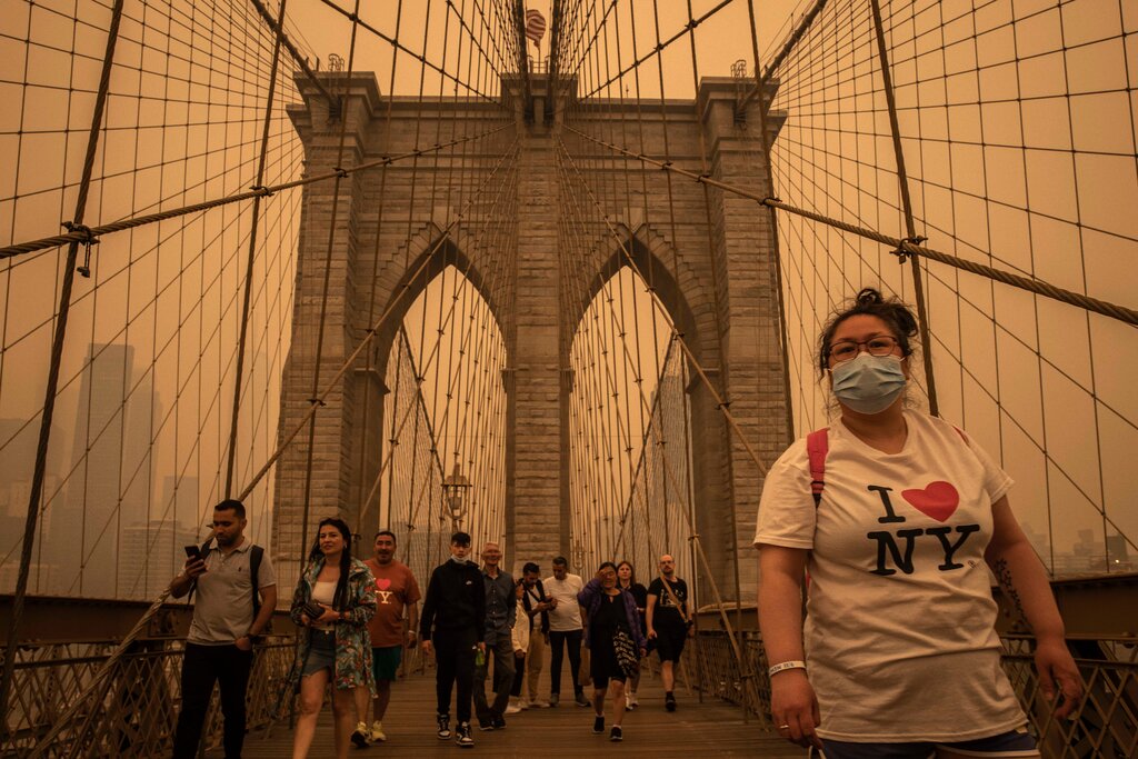 A woman in a shirt reading “I Heart NY” and wearing a blue mask crosses the Brooklyn Bridge, as the sky behind her glows a hazy orange.