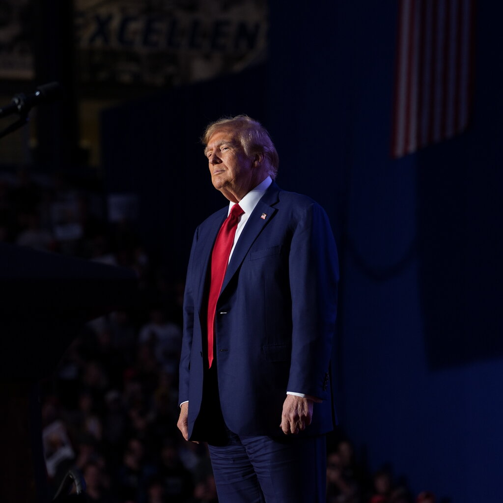 Donald Trump, wearing a blue suit and red tie, stands on a stage. An audience is visible behind him in the shadows.