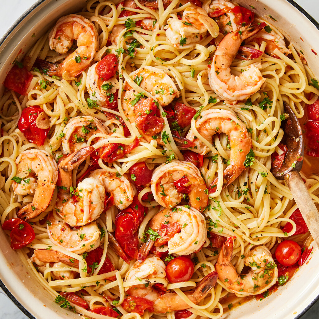 A white Dutch oven holds shrimp pasta with burst cherry tomatoes and a sprinkling of chopped parsley.