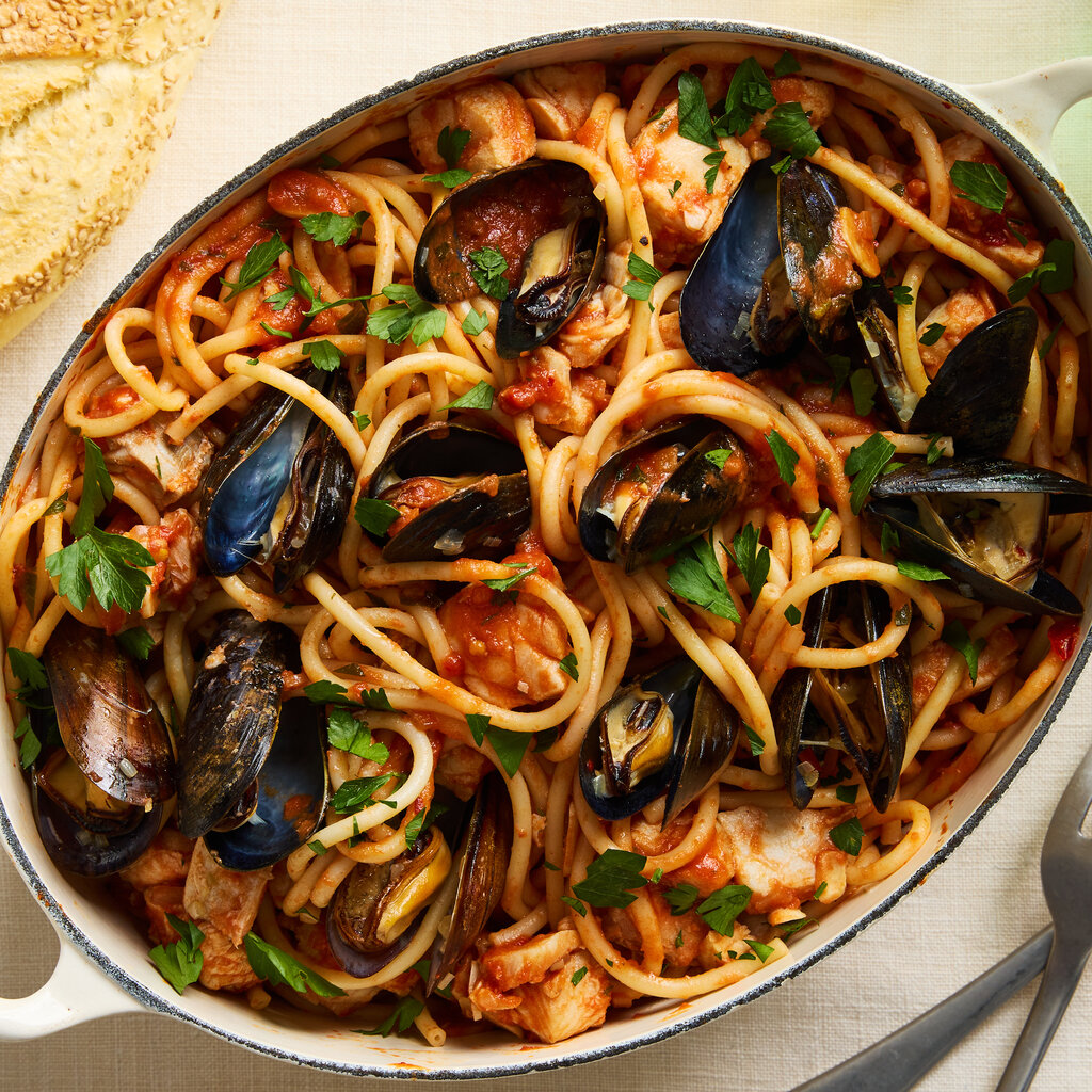 An oval Dutch oven holds bucatini with mussels and cod in a tomato sauce, with serving utensils and a loaf of seeded bread nearby.
