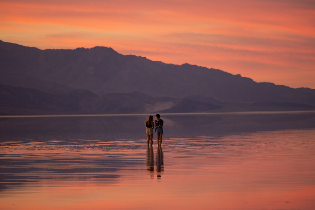 Two people standing in a lake at sunset. The sky is orange and pink and reflecting off the water's surface. Mountains are in the distance.