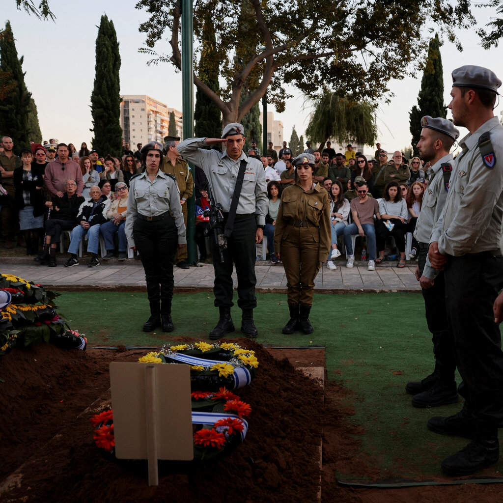 A military officer stands at attention in front of a grave covered in a mound of dirt with a wreath of flowers on top. Other officers stand round the grave, and there is a large group of other attendees behind.