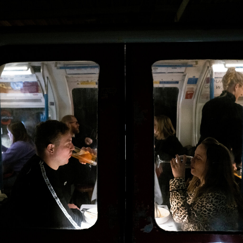 A view from the outside of two people dining inside a Tube train car. A man and a woman sitting opposite each other sip from glassed, other diners can be seen in background. 