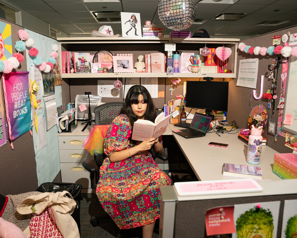 A woman reads a book in a cubicle covered in pink and blue decorations with a disco ball hanging from the ceiling.