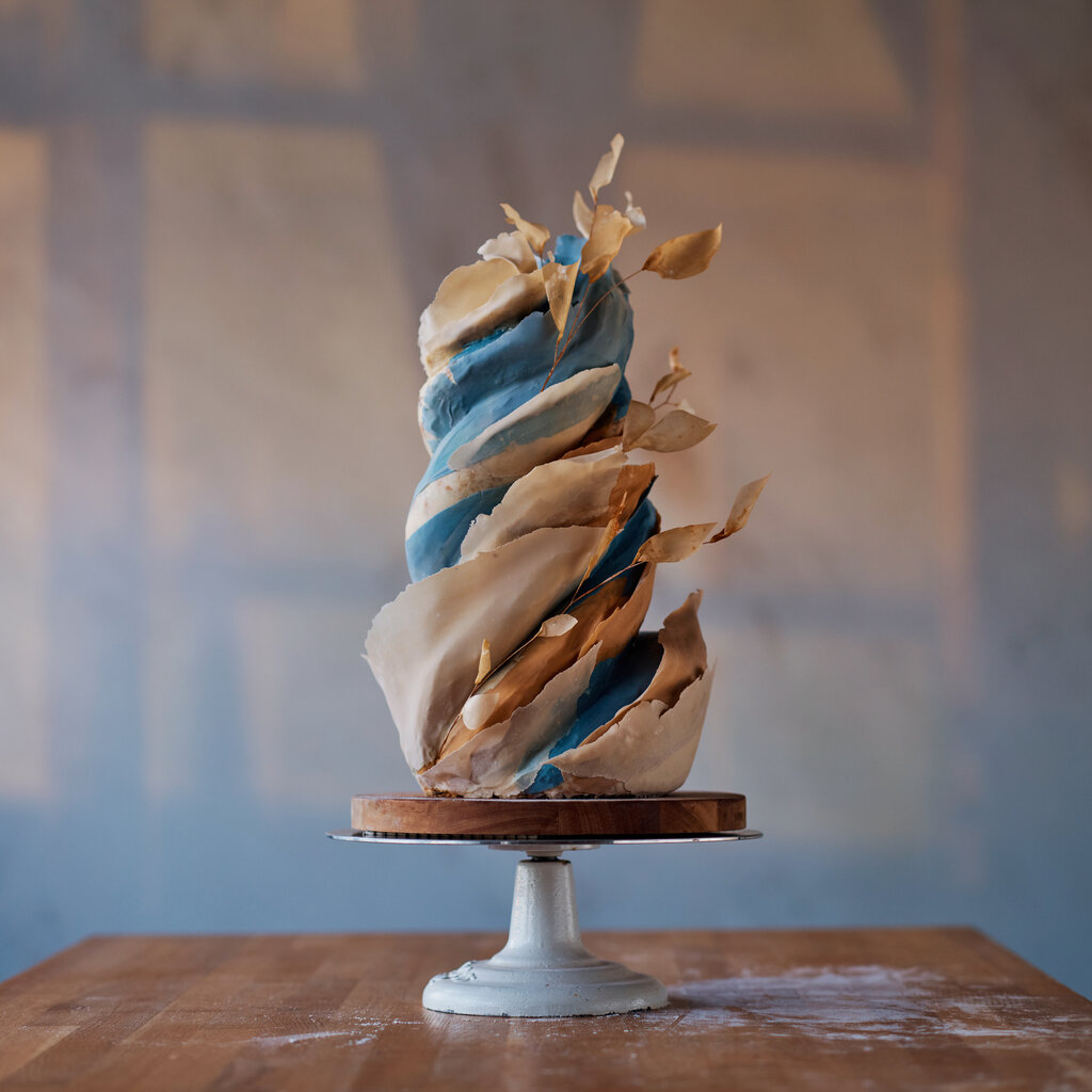 The finished product: a cake, colored in blue and shades of beige, with “flowers” jutting out of it, on a cake stand and a partially floured table. Faded light from nearby windows color the wall behind the scene. 