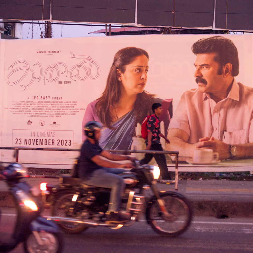 People on foot and motorcycle pass by a large movie poster featuring a man and a woman looking at each other.