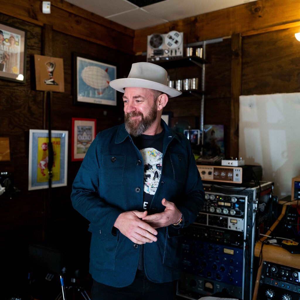 Kristian Bush, wearing a hat, looks away and smiles as he stands by musical instruments and amplifiers. His blue jacket covers a black T-shirt with a white skull design on it. 