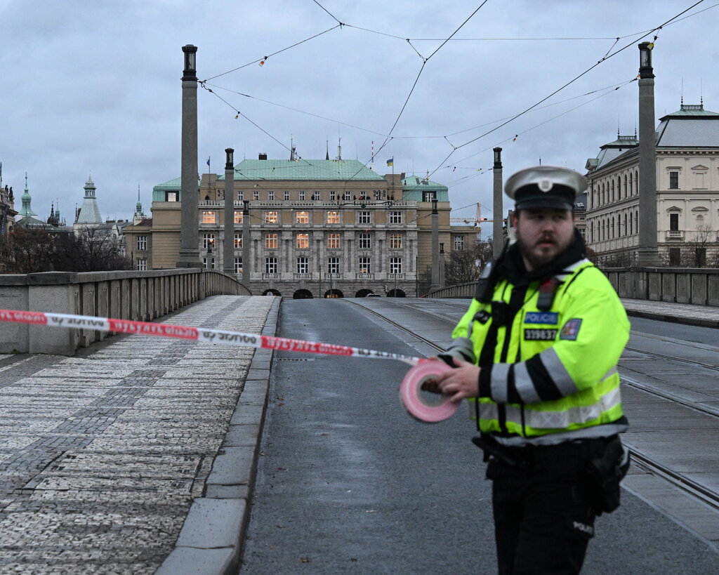 A police officer rolls crime tape across a road leading to a large stone university building on the other side of a bridge.